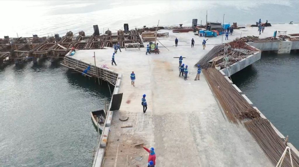 A construction site in Palawan shows multiple workers assembling rebar frameworks and concrete sections on a partially built pier extending over calm water, representing active infrastructure development supporting Palawan property investment.