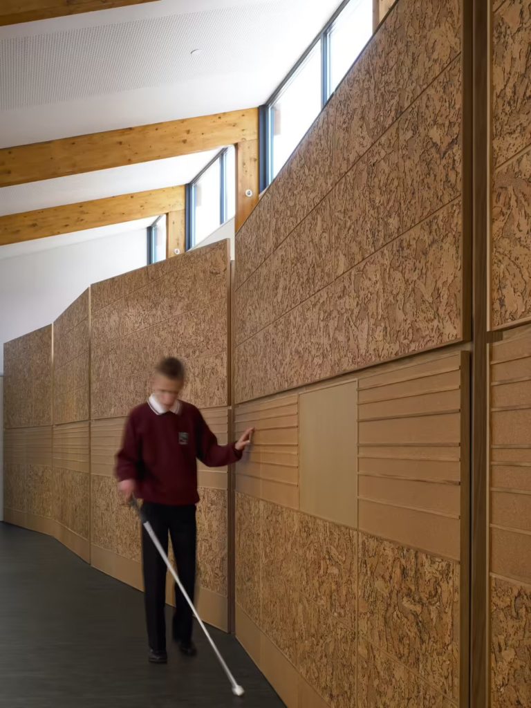 A young boy walks alongside an indoor corridor wall finished in tactile stone and timber textures, lit by diffused natural light through clerestory windows, within Hazelwood School in Glasgow designed for children with multiple sensory impairments.
