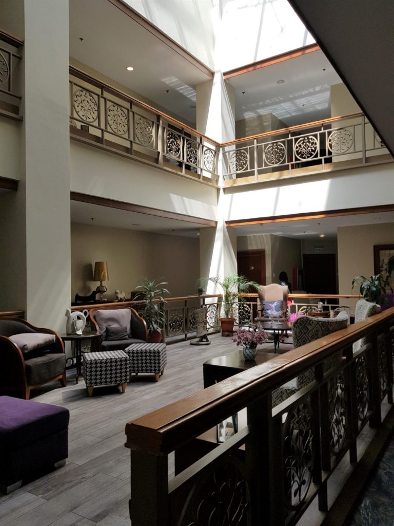 A spacious multi-level hotel atrium features sunlit balconies, large windows, natural finishes, and classic decorative elements, illustrating sustainable interior design that prioritizes passive energy strategies and environmental responsiveness.