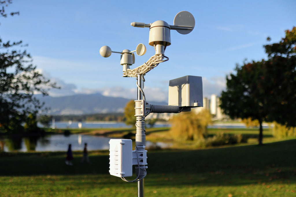 A weather station with a tall sensor mast and surrounding instrumentation set within a landscaped park, illustrating environmental monitoring infrastructure used in sustainable urban waste management systems.