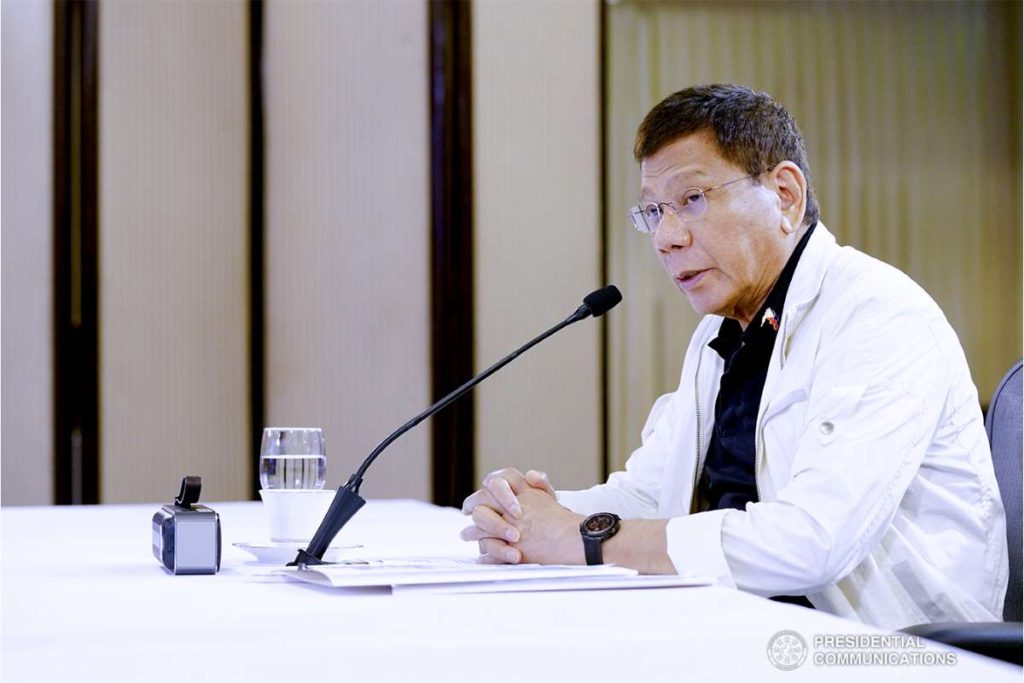 President Rodrigo Roa Duterte seated at a table speaking into a microphone, wearing a white long-sleeved shirt over a dark inner top, in a formal conference or press briefing room with a minimal table and microphone setup, conveying a serious expression while delivering a key policy statement on Philippine energy strategy.