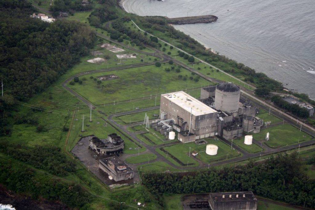 Aerial view of the Bataan Nuclear Power Plant showing its full facility complex, including reactor building, adjacent cooling systems, and support structures, situated near coastal terrain with surrounding green slopes, captured under clear skies to visually represent the integration of nuclear infrastructure in the Philippine energy sector.