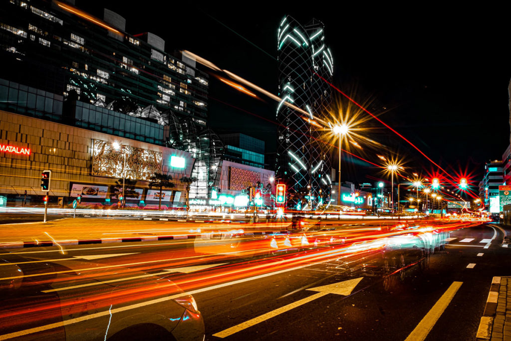 A nighttime city street illuminated by vehicle headlights and traffic signals, with multiple cars in motion and buildings in the background, representing an advanced urban environment utilizing real-time traffic control systems.